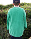 A boy viewed from behind wearing a bright green corded crewneck long-sleeve sweatshirt and a dark baseball cap, standing outdoors in front of green bushes.