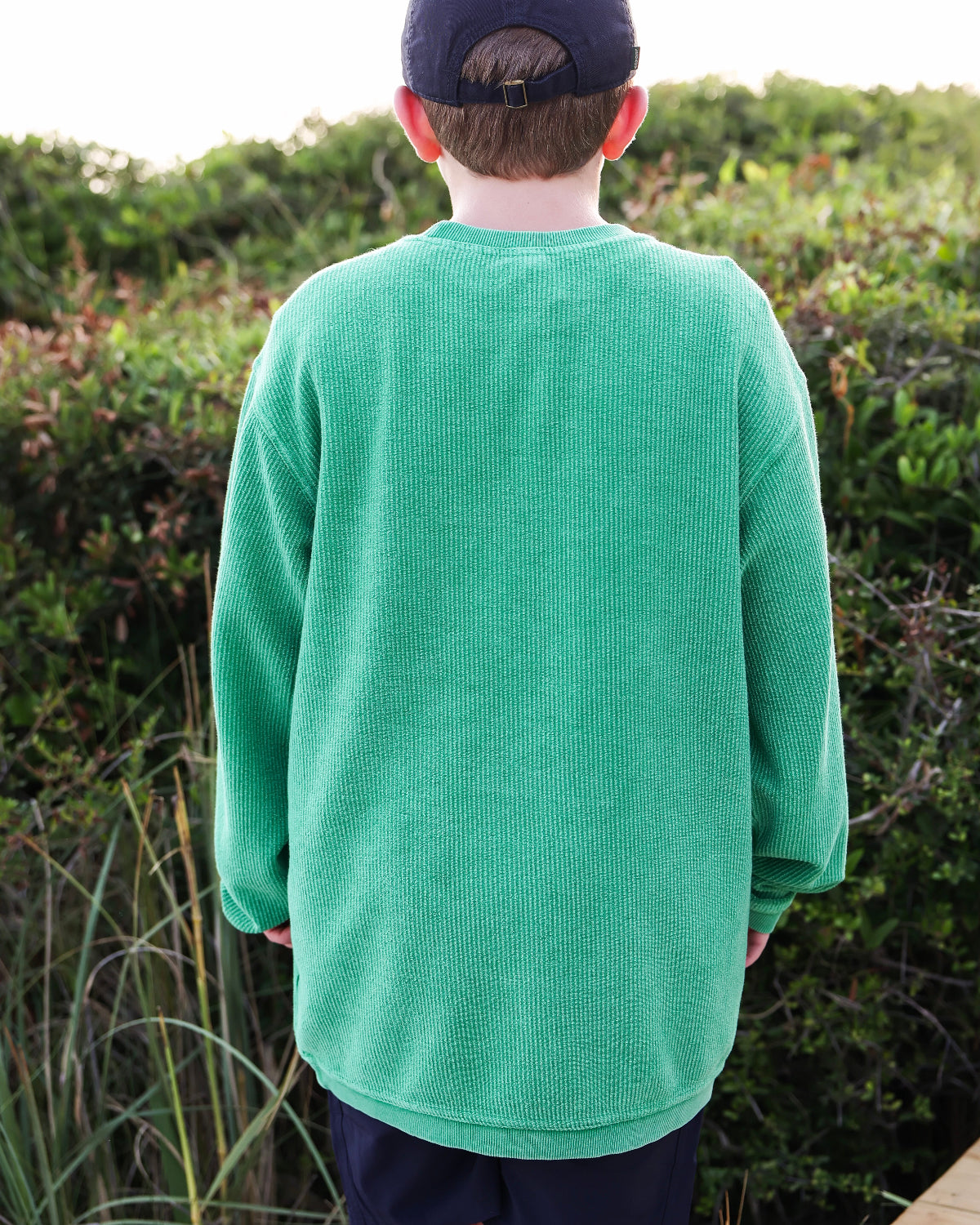 A boy viewed from behind wearing a bright green corded crewneck long-sleeve sweatshirt and a dark baseball cap, standing outdoors in front of green bushes.
