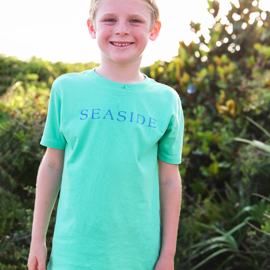 A smiling young boy wearing a clover green short-sleeve t-shirt with 'SEASIDE' printed in blue across the chest, standing outdoors in front of green foliage.