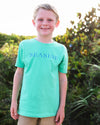 A smiling young boy wearing a clover green short-sleeve t-shirt with 'SEASIDE' printed in blue across the chest, standing outdoors in front of green foliage.