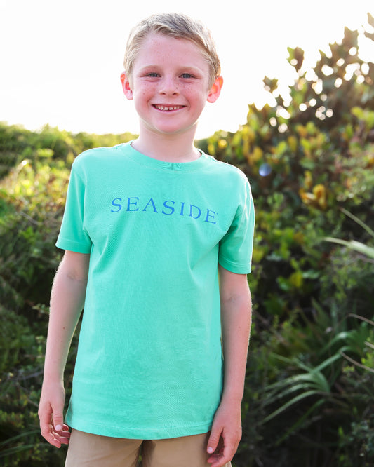 A smiling young boy wearing a clover green short-sleeve t-shirt with 'SEASIDE' printed in blue across the chest, standing outdoors in front of green foliage.