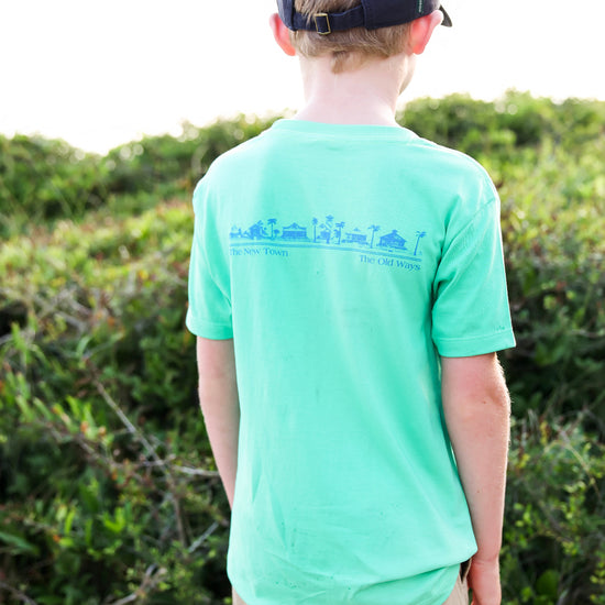 A child viewed from behind wearing a mint green t-shirt with a blue graphic print on the back, paired with a navy baseball cap and khaki pants, standing outdoors in front of green bushes.