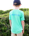 A child viewed from behind wearing a mint green t-shirt with a blue graphic print on the back, paired with a navy baseball cap and khaki pants, standing outdoors in front of green bushes.