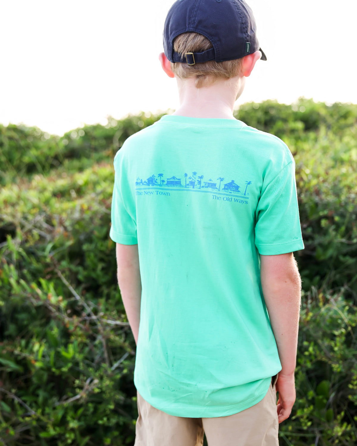A child viewed from behind wearing a mint green t-shirt with a blue graphic print on the back, paired with a navy baseball cap and khaki pants, standing outdoors in front of green bushes.