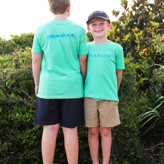 Two boys standing barefoot on a wooden deck outdoors, wearing matching clover green t-shirts. The boy on the left has his back to the camera showing a blue graphic on the back, and the boy on the right is facing forward smiling, wearing a black cap and khaki shorts.