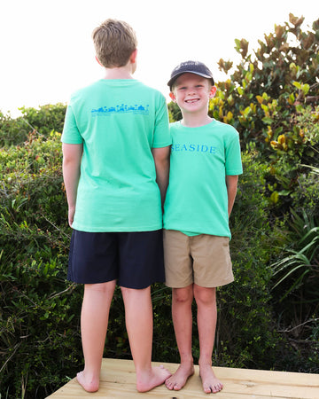 Two boys standing barefoot on a wooden deck outdoors, wearing matching clover green t-shirts. The boy on the left has his back to the camera showing a blue graphic on the back, and the boy on the right is facing forward smiling, wearing a black cap and khaki shorts.