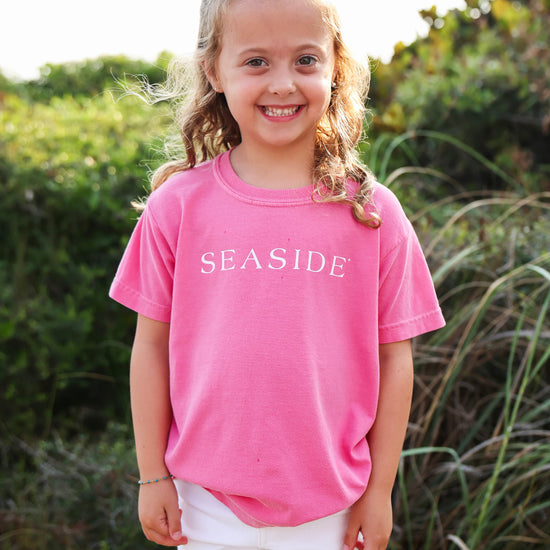 A young girl with blonde braids stands outdoors wearing a bright pink t-shirt with 'SEASIDE' printed in white across the chest, paired with white shorts.