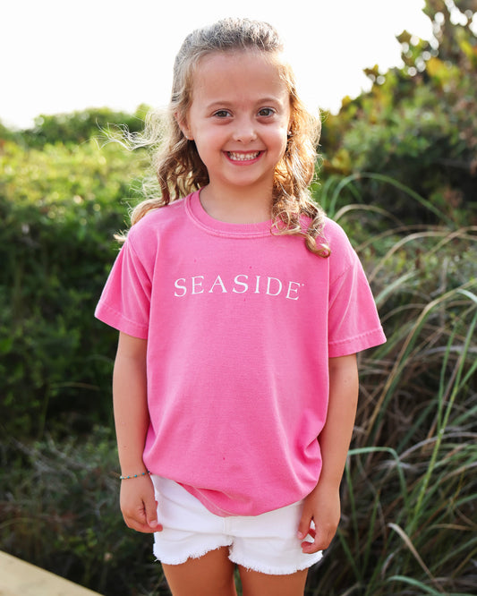 A young girl with blonde braids stands outdoors wearing a bright pink t-shirt with 'SEASIDE' printed in white across the chest, paired with white shorts.