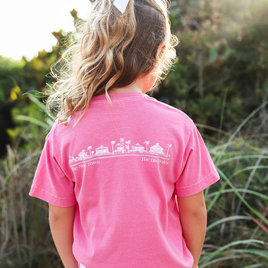 A young girl viewed from behind wearing a bright pink t-shirt with a white graphic print of a beach town and the words 'new town' and 'the old ways'. She is standing outdoors in a grassy field with soft-focus green foliage and warm sunlight in the background.