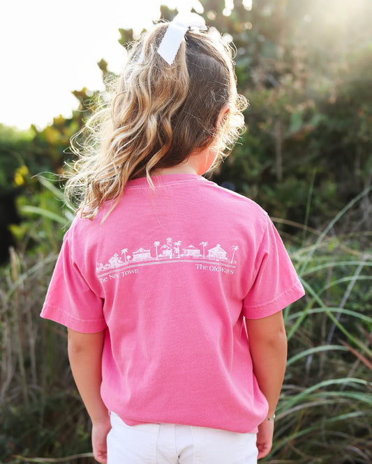 A young girl viewed from behind wearing a bright pink t-shirt with a white graphic print of a beach town and the words 'new town' and 'the old ways'. She is standing outdoors in a grassy field with soft-focus green foliage and warm sunlight in the background.