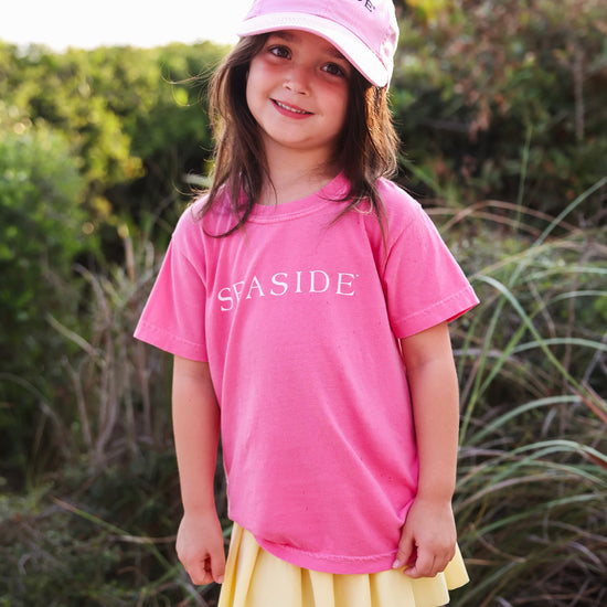 A young girl wearing a pink short-sleeve t-shirt with 'SEASIDE' printed in white across the chest, a matching pink baseball cap, and a yellow pleated skirt, standing outdoors in front of green foliage.