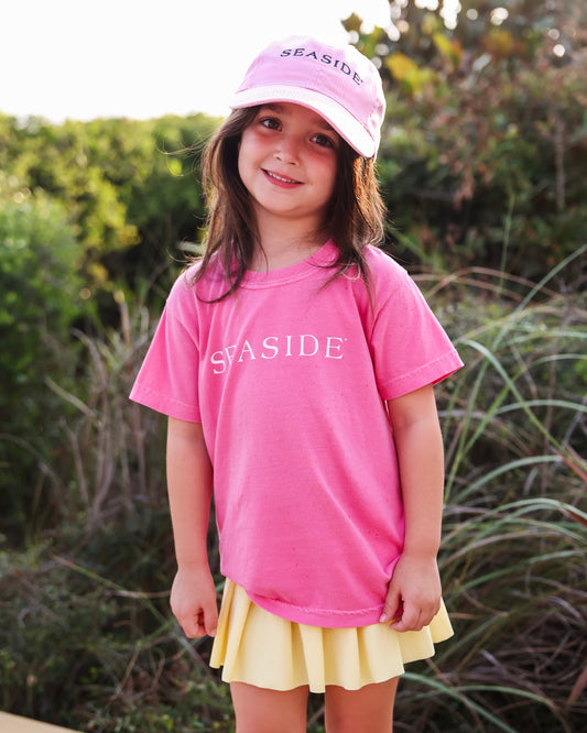 A young girl wearing a pink short-sleeve t-shirt with 'SEASIDE' printed in white across the chest, a matching pink baseball cap, and a yellow pleated skirt, standing outdoors in front of green foliage.