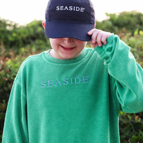 A child wearing a green ribbed knit long-sleeve shirt with 'SEASIDE' embroidered on the chest and a navy baseball cap with 'SEASIDE' embroidered on the front, standing outdoors in front of a soft-focus green background.