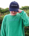 A child wearing a green ribbed knit long-sleeve shirt with 'SEASIDE' embroidered on the chest and a navy baseball cap with 'SEASIDE' embroidered on the front, standing outdoors in front of a soft-focus green background.