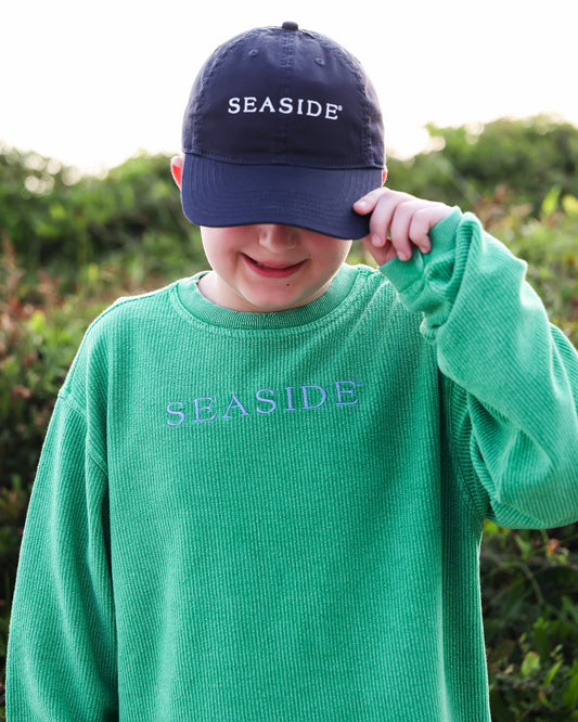 A child wearing a green ribbed knit long-sleeve shirt with 'SEASIDE' embroidered on the chest and a navy baseball cap with 'SEASIDE' embroidered on the front, standing outdoors in front of a soft-focus green background.