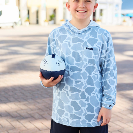 Child wearing a blue patterned hoodie and cap, holding a football with 'Seaside' branding.