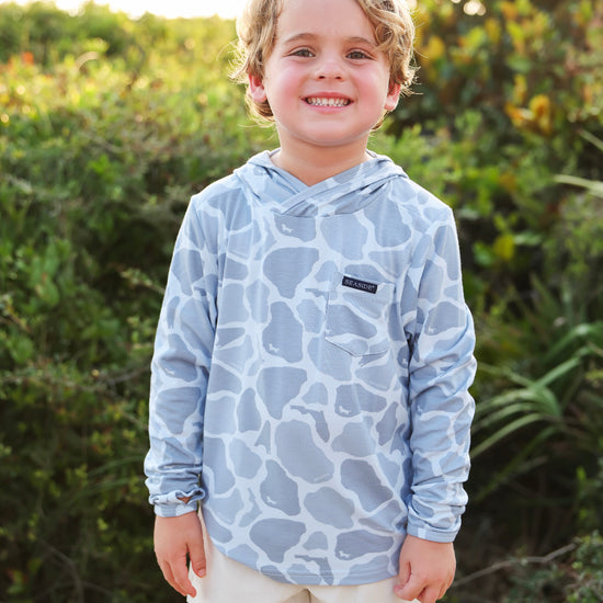 A smiling young boy wearing a light blue and white camo-print long-sleeve hooded shirt and off-white shorts, standing outdoors in front of a blurred green foliage background.
