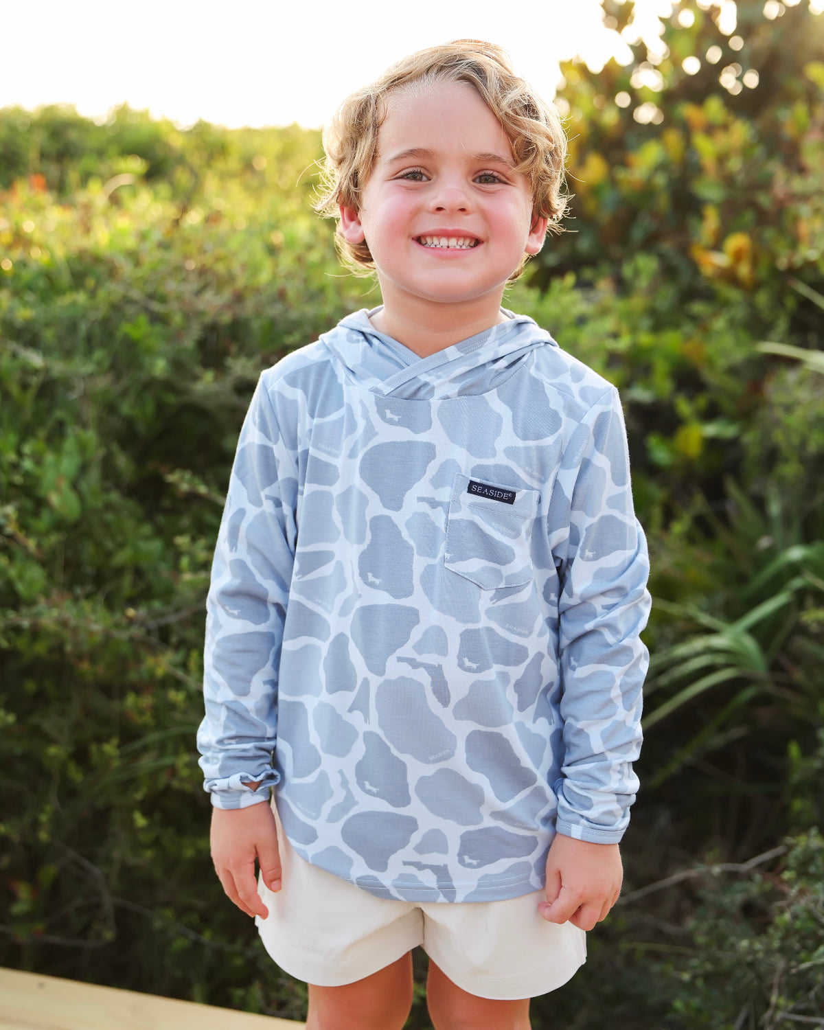 A smiling young boy wearing a light blue and white camo-print long-sleeve hooded shirt and off-white shorts, standing outdoors in front of a blurred green foliage background.