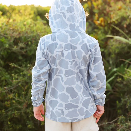 A child viewed from behind wearing a light blue and white camo-print hooded sun hoodie and beige shorts, standing outdoors in front of a blurred green bush background.