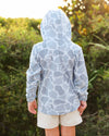 A child viewed from behind wearing a light blue and white camo-print hooded sun hoodie and beige shorts, standing outdoors in front of a blurred green bush background.