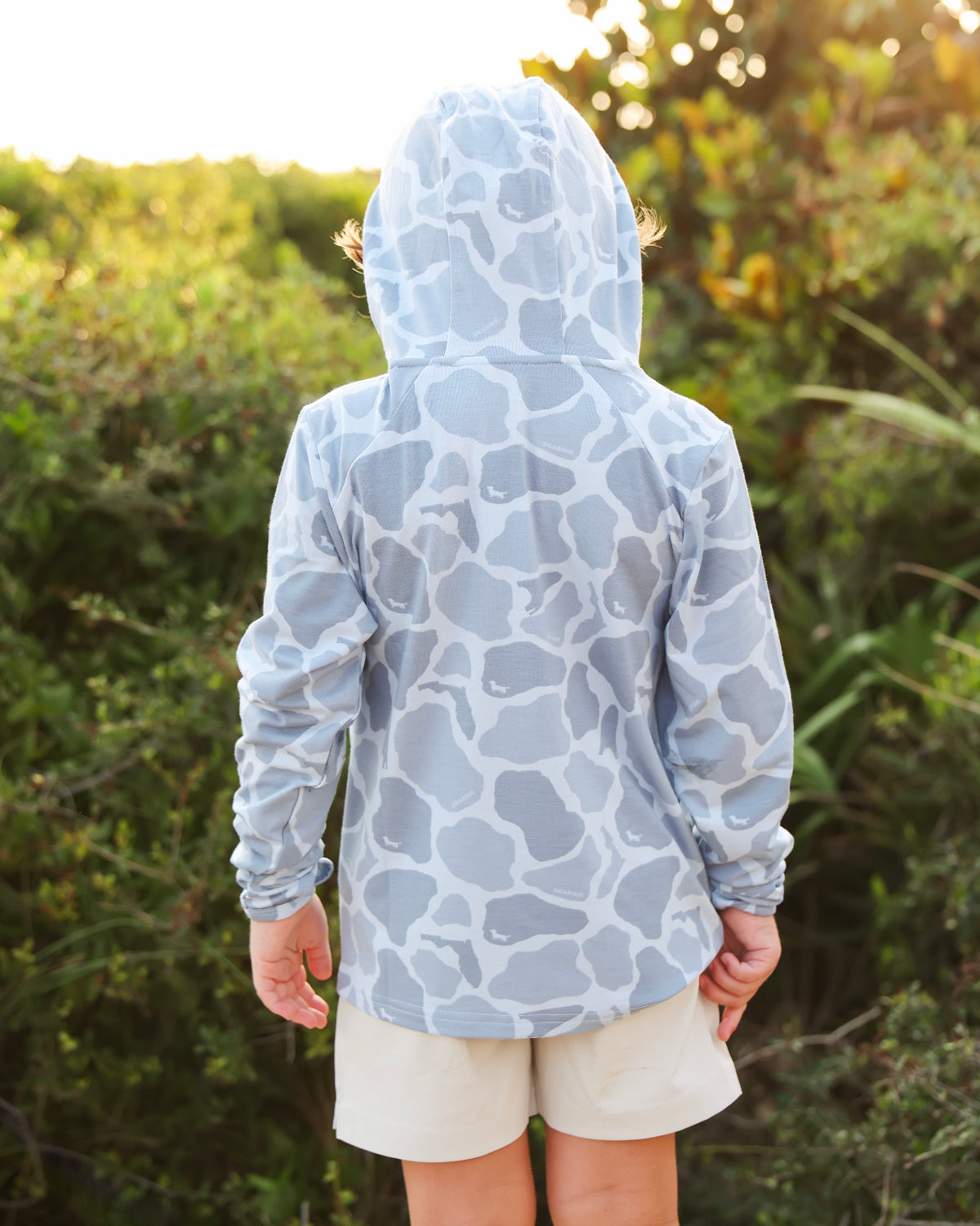 A child viewed from behind wearing a light blue and white camo-print hooded sun hoodie and beige shorts, standing outdoors in front of a blurred green bush background.