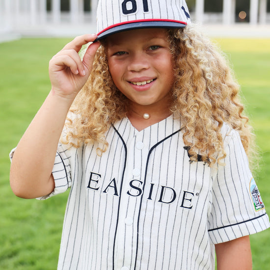 A young girl with long curly blonde hair wearing a white pinstripe baseball jersey and matching cap with the number 81, standing outdoors on a grassy field. The jersey has 'SEASIDE' printed across the chest.