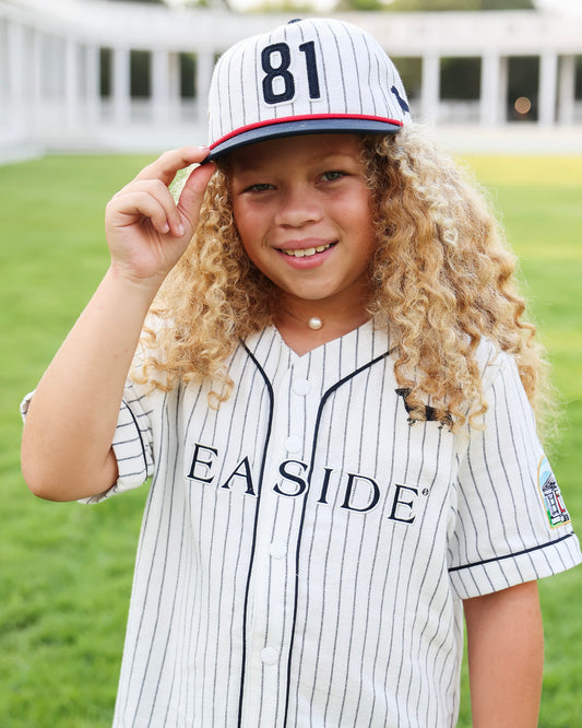 A young girl with long curly blonde hair wearing a white pinstripe baseball jersey and matching cap with the number 81, standing outdoors on a grassy field. The jersey has 'SEASIDE' printed across the chest.
