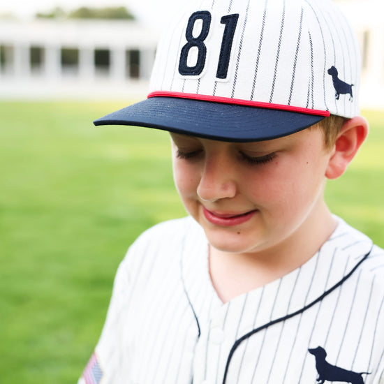 A young boy wearing a white pinstripe baseball cap with navy blue trim, the number 81 on the front, and a small dachshund logo, paired with a matching white pinstripe baseball jersey. The background is a soft-focus green grass field.