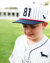 A young boy wearing a white pinstripe baseball cap with navy blue trim, the number 81 on the front, and a small dachshund logo, paired with a matching white pinstripe baseball jersey. The background is a soft-focus green grass field.