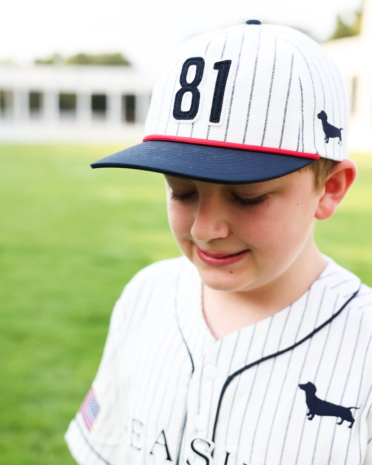 A young boy wearing a white pinstripe baseball cap with navy blue trim, the number 81 on the front, and a small dachshund logo, paired with a matching white pinstripe baseball jersey. The background is a soft-focus green grass field.