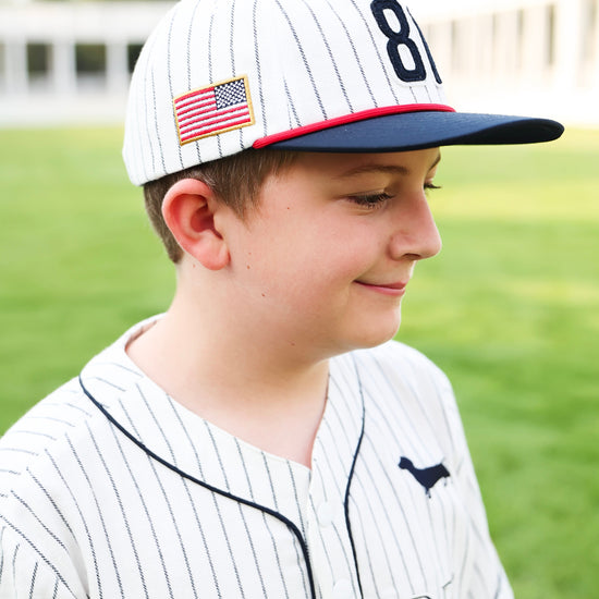 A young boy wearing a white pinstripe baseball cap with a navy blue brim, a navy number 81, and an American flag patch, paired with a matching white pinstripe baseball jersey on a blurred green field background.