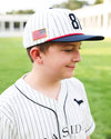 A young boy wearing a white pinstripe baseball cap with a navy blue brim, a navy number 81, and an American flag patch, paired with a matching white pinstripe baseball jersey on a blurred green field background.