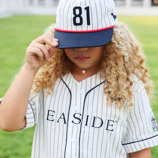 A young girl with long curly blonde hair wearing a white pinstripe baseball jersey and matching cap with the number 81. The cap has a navy blue brim with a thin red stripe, and the jersey features the word 'SEASIDE' across the chest.