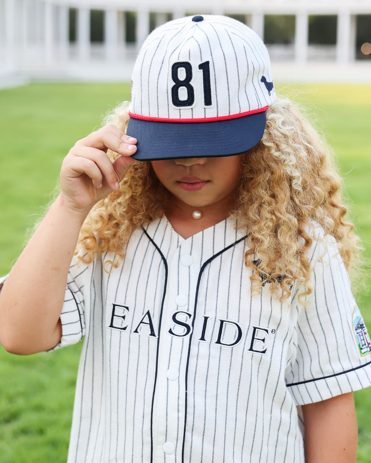 A young girl with long curly blonde hair wearing a white pinstripe baseball jersey and matching cap with the number 81. The cap has a navy blue brim with a thin red stripe, and the jersey features the word 'SEASIDE' across the chest.
