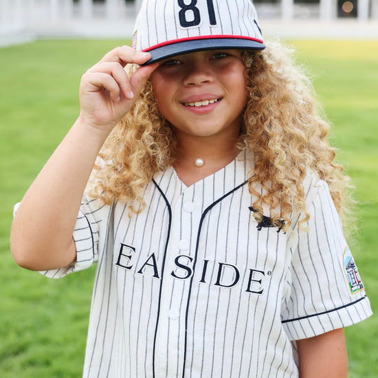 A young girl with long curly blonde hair wearing a white pinstripe baseball jersey and matching cap with the number 81, standing outdoors on a green lawn with a building in the background.