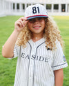 A young girl with long curly blonde hair wearing a white pinstripe baseball jersey and matching cap with the number 81, standing outdoors on a green lawn with a building in the background.