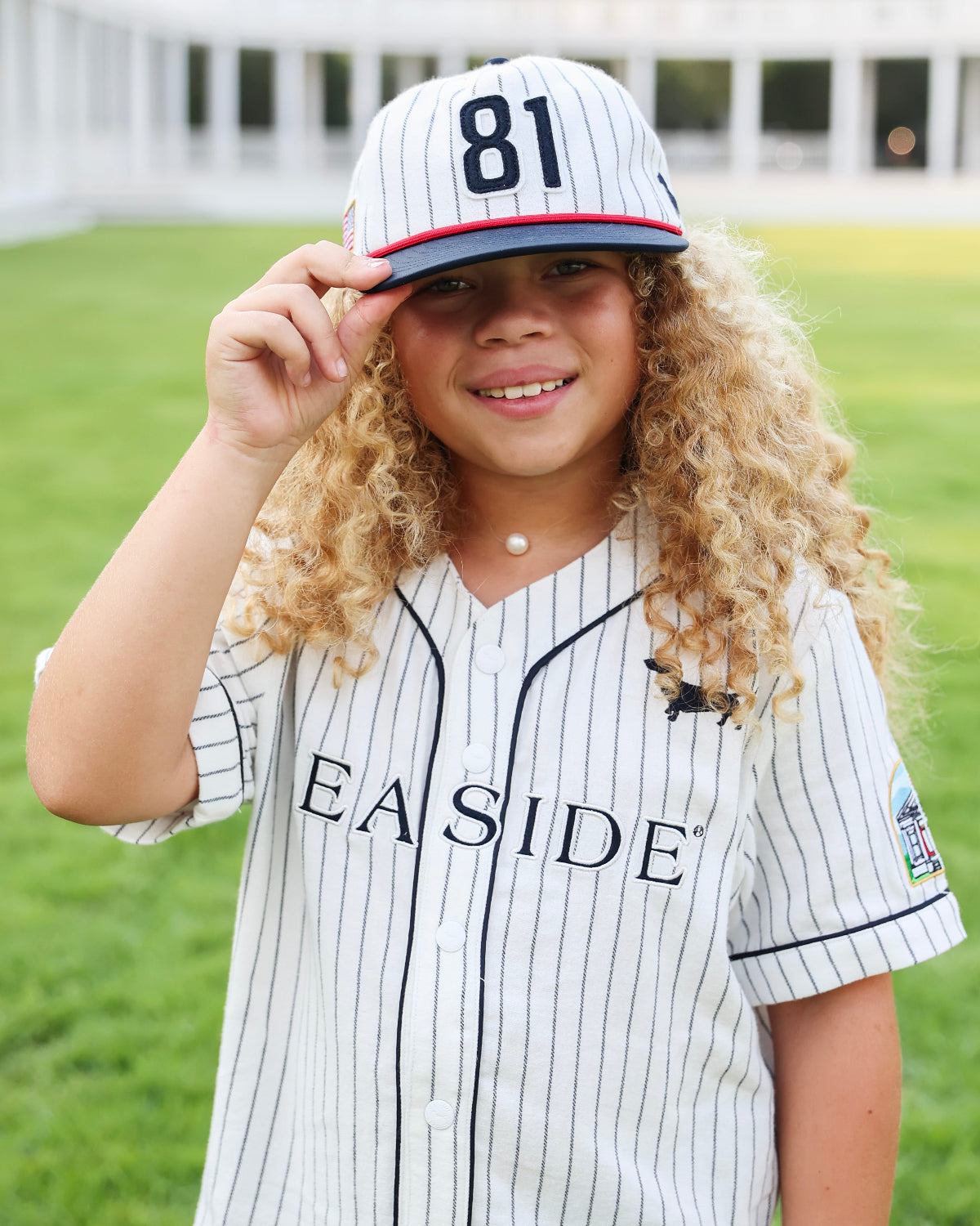 A young girl with long curly blonde hair wearing a white pinstripe baseball jersey and matching cap with the number 81, standing outdoors on a green lawn with a building in the background.