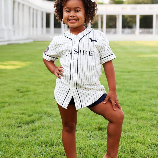 A young girl with curly hair stands barefoot on a green lawn wearing a short baseball-style jersey with 'SEASIDE' printed across the front, navy shorts, and a white headband.