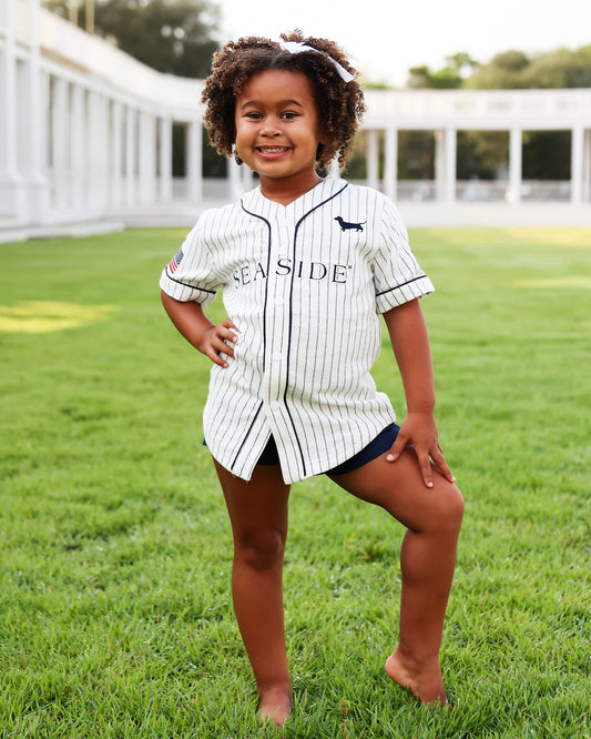 A young girl with curly hair stands barefoot on a green lawn wearing a short baseball-style jersey with 'SEASIDE' printed across the front, navy shorts, and a white headband.