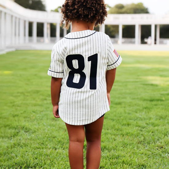 A child standing on a green lawn, viewed from behind, wearing a white pinstripe baseball-style shirt with the number 81 on the back.