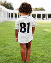 A child standing on a green lawn, viewed from behind, wearing a white pinstripe baseball-style shirt with the number 81 on the back.