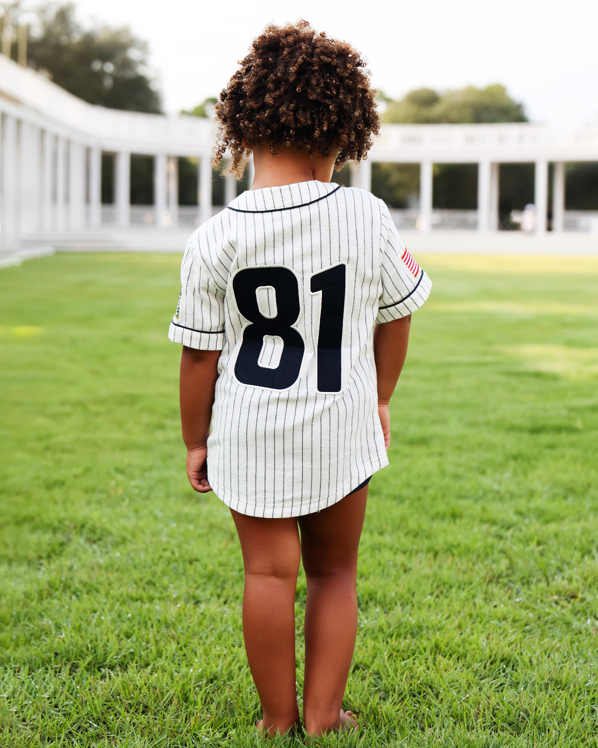 A child standing on a green lawn, viewed from behind, wearing a white pinstripe baseball-style shirt with the number 81 on the back.