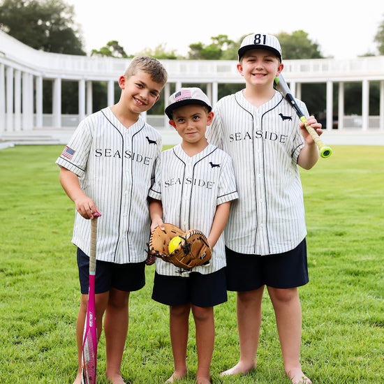Three boys wearing matching pinstriped Seaside baseball jerseys, navy shorts, and baseball caps stand barefoot on a grassy field, each holding a baseball bat or glove.