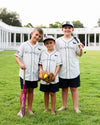 Three boys wearing matching pinstriped Seaside baseball jerseys, navy shorts, and baseball caps stand barefoot on a grassy field, each holding a baseball bat or glove.