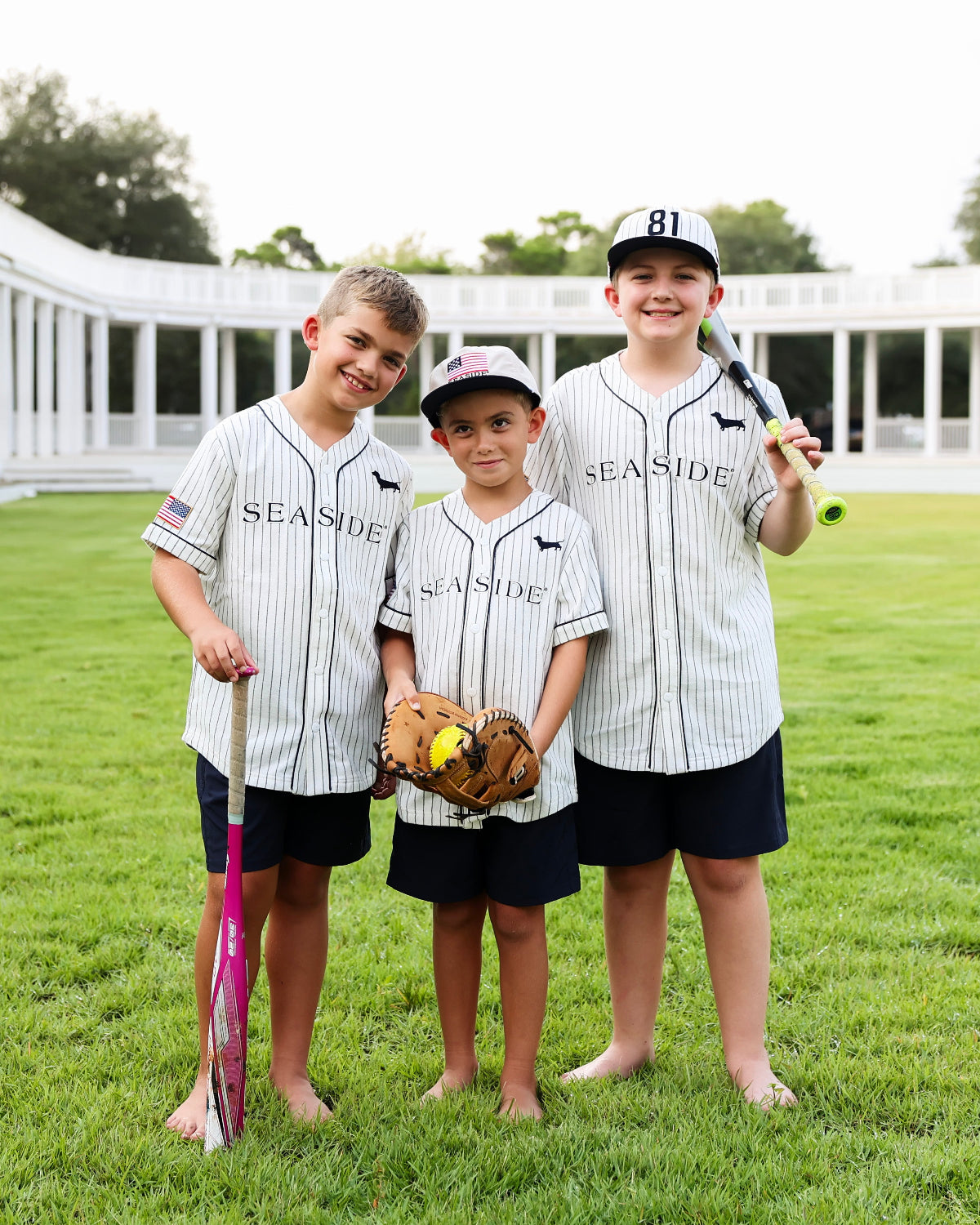 Three boys wearing matching pinstriped Seaside baseball jerseys, navy shorts, and baseball caps stand barefoot on a grassy field, each holding a baseball bat or glove.