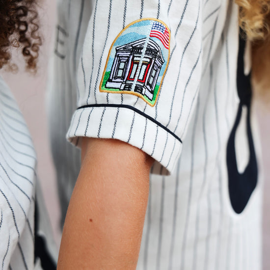 Close-up of a person wearing a white pinstripe baseball jersey with a colorful embroidered patch on the sleeve of the seaside post office.