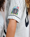 Close-up of a person wearing a white pinstripe baseball jersey with a colorful embroidered patch on the sleeve of the seaside post office.
