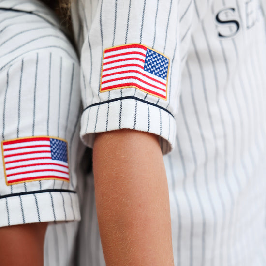 Close-up of a person wearing a white pinstriped baseball-style shirt with rolled sleeves and American flag patches on both upper arms.