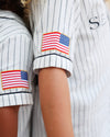 Close-up of a person wearing a white pinstriped baseball-style shirt with rolled sleeves and American flag patches on both upper arms.