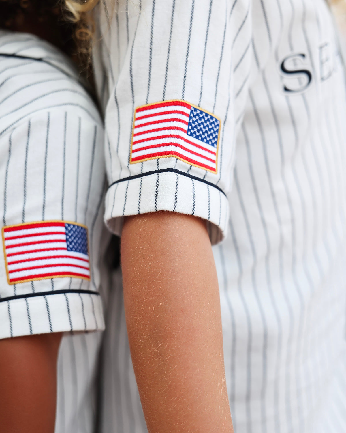 Close-up of a person wearing a white pinstriped baseball-style shirt with rolled sleeves and American flag patches on both upper arms.